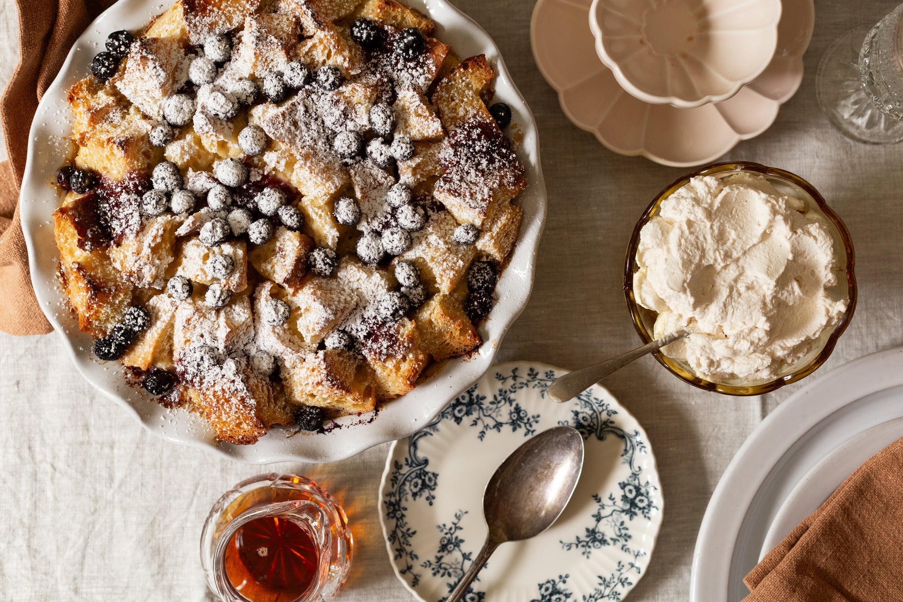 Baked pudding with berries and cream on a table with plates and a spoon.