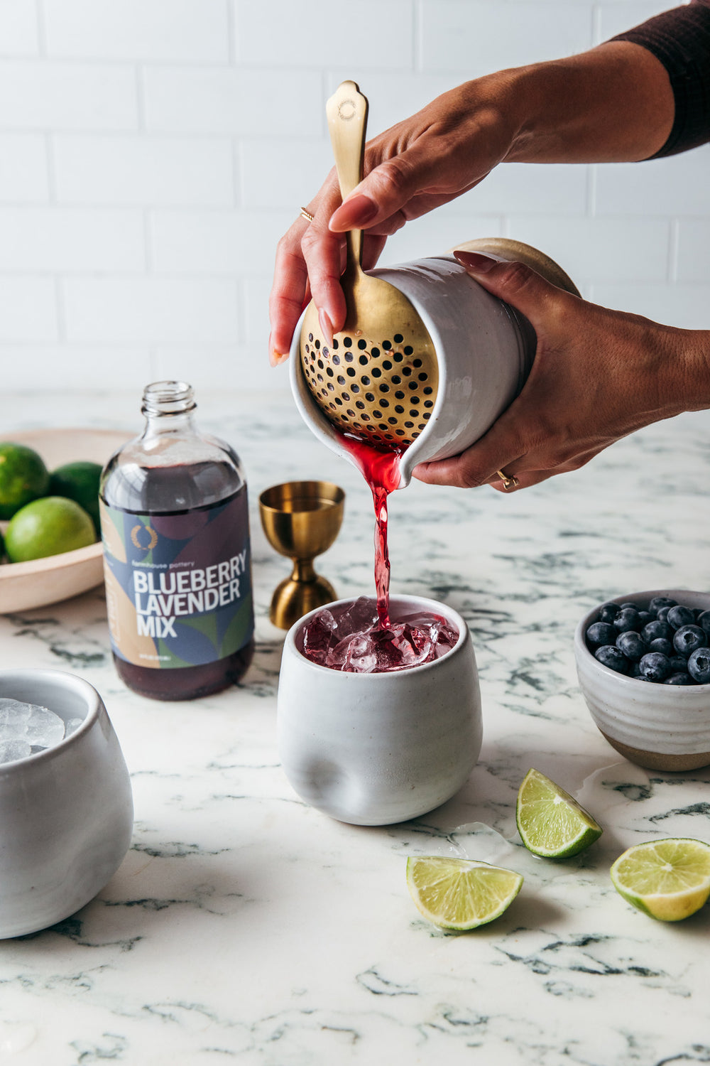 Person pouring a red liquid from a strainer into a white bowl on a marble surface with ingredients and a bottle labeled 'Blueberry Lavender Mix'.